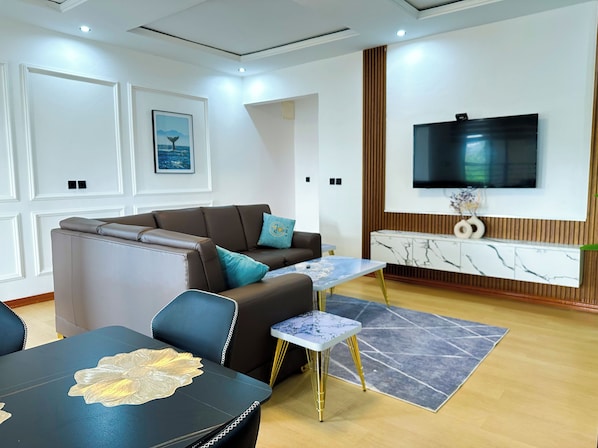 A modern living room with a sectional sofa, a TV mounted on the wall, a marble console table, and a dining area with a table and chairs.