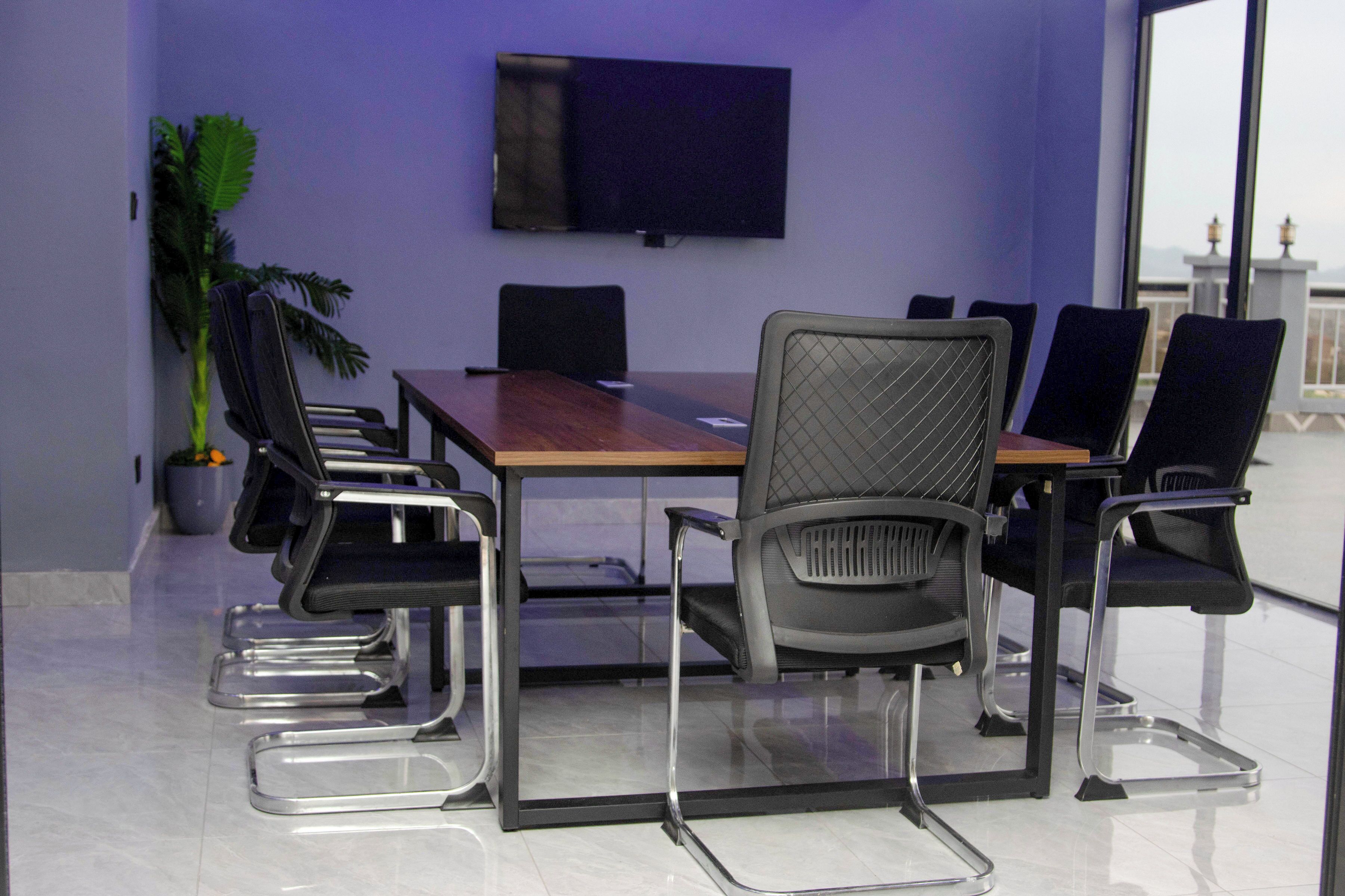 A conference room with a wooden table, black chairs, a large screen, and a potted plant.