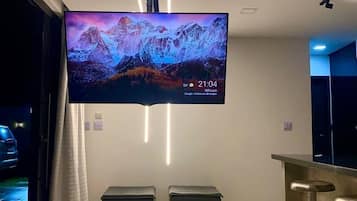 Una sala de estar moderna con un televisor de pantalla plana grande montado en la pared, una zona de bar y un espacio de estar con otomanas.
