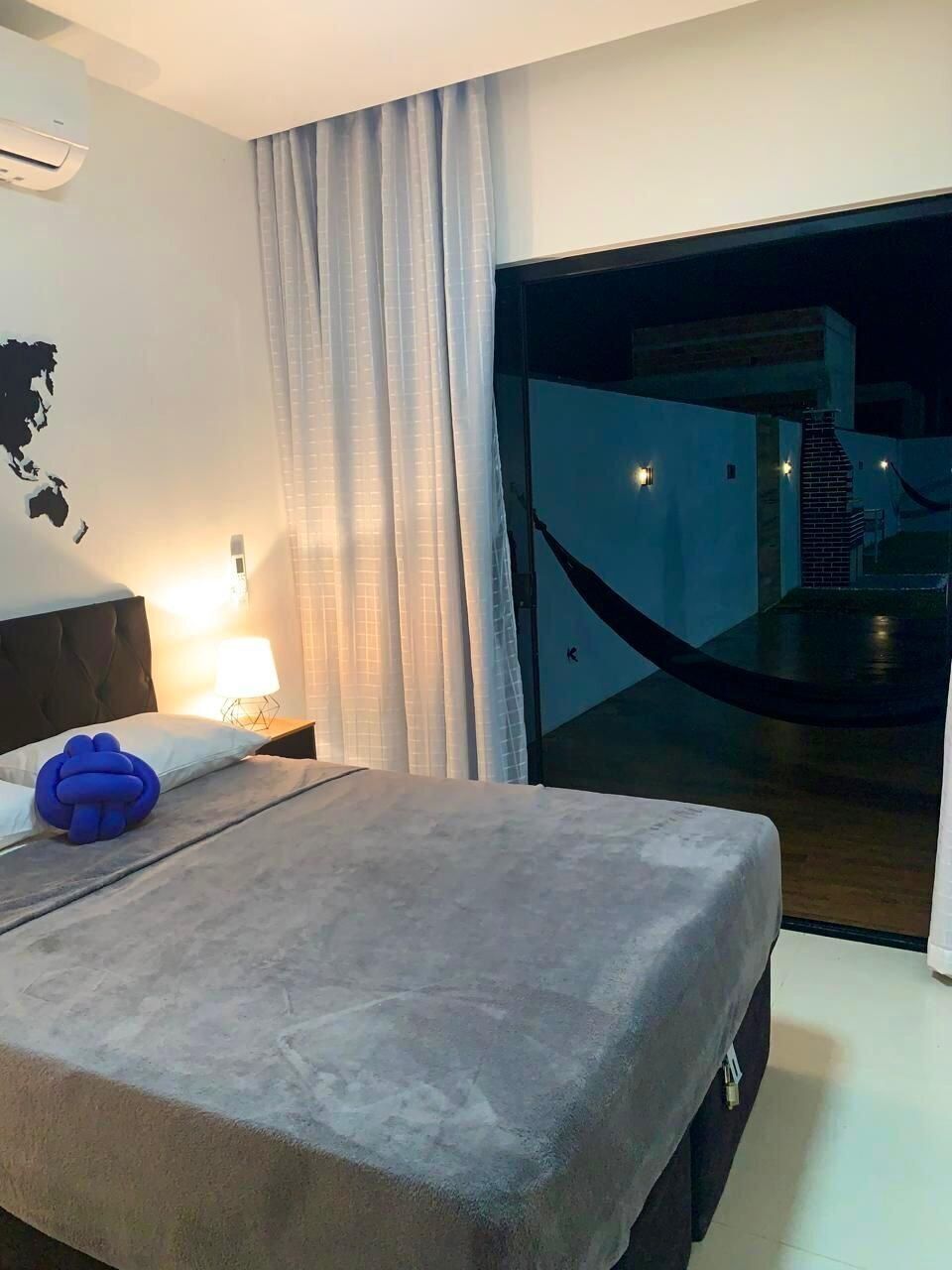 A hotel room with a bed, a nightstand, a lamp, a world map on the wall, and a view of the outside through a glass door.