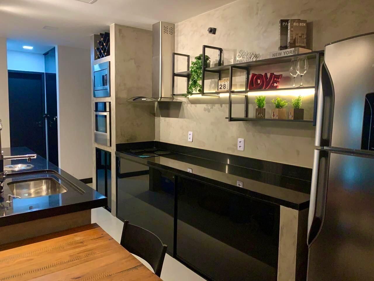 Una cocina moderna con un refrigerador de acero inoxidable, un horno empotrado y una encimera negra. Hay una mesa de comedor de madera y una repisa con objetos decorativos.