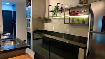 Una cocina moderna con un refrigerador de acero inoxidable, un horno empotrado y una encimera negra. Hay una mesa de comedor de madera y una repisa con objetos decorativos.