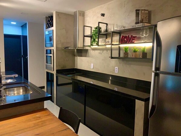 A modern kitchen with a stainless steel refrigerator, a built-in oven, and a black countertop. There is a wooden dining table and a shelf with decorative items.