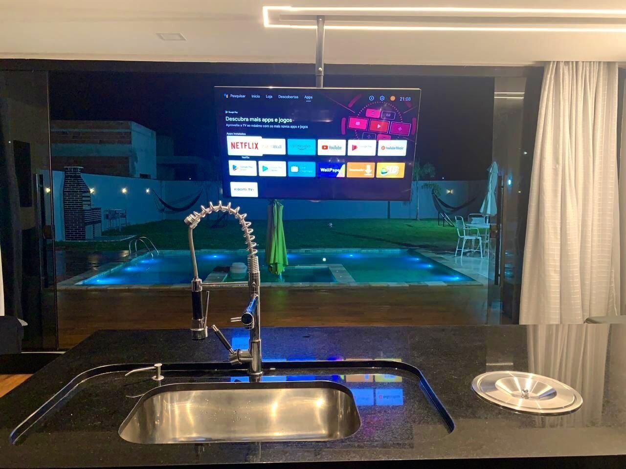 Uma cozinha moderna com pia de aço inoxidável, uma TV de tela plana montada acima e vista para uma piscina lá fora.
