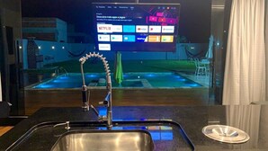 A modern kitchen with a stainless steel sink, a flat-screen TV mounted above it, and a view of a swimming pool outside. - CASA PREMIUM - ARRAIAL DO CABO - RJ (Arraial do Cabo)