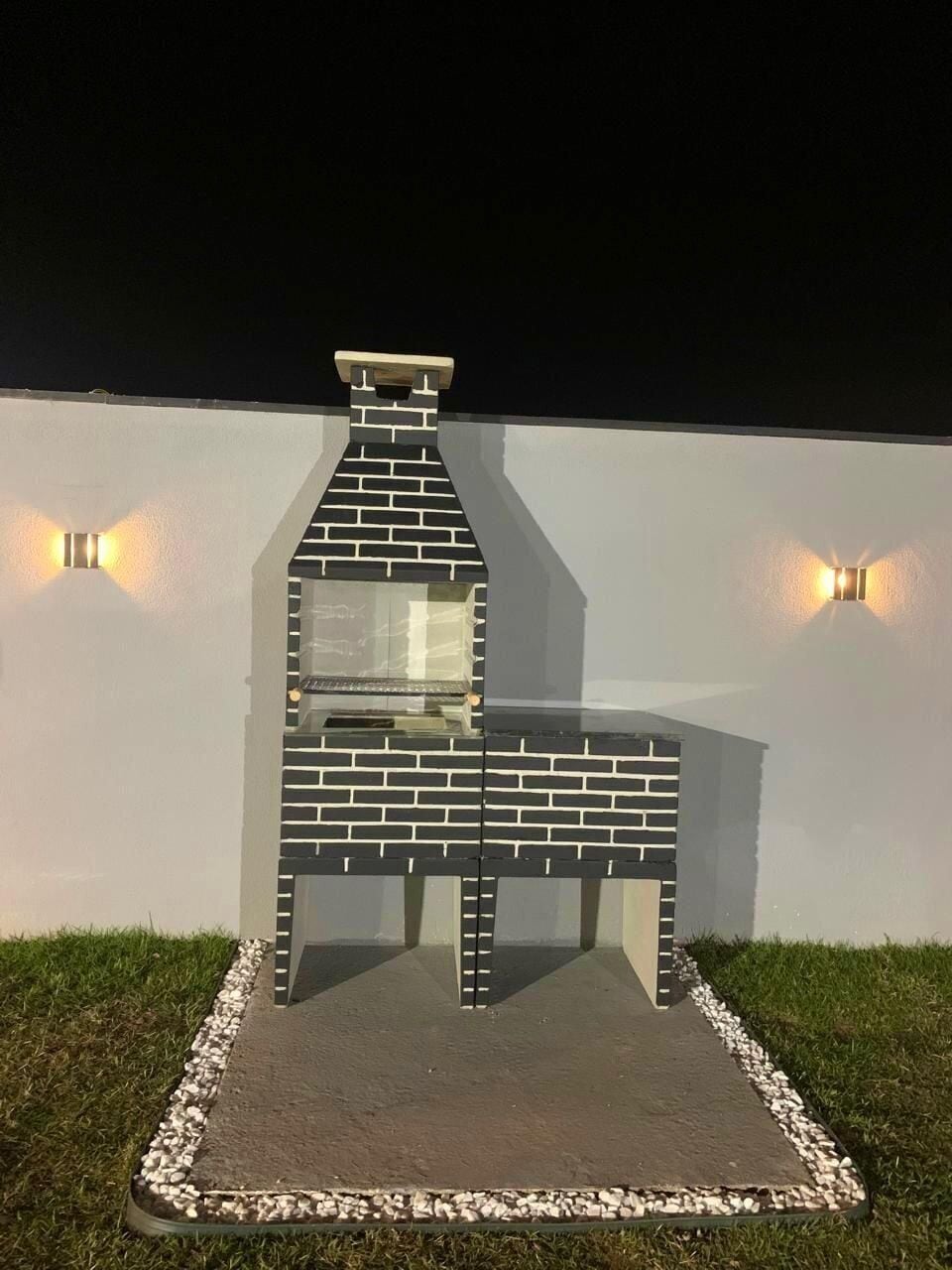 Una parrilla de ladrillo con chimenea, frente a una pared blanca con dos luces empotradas.
