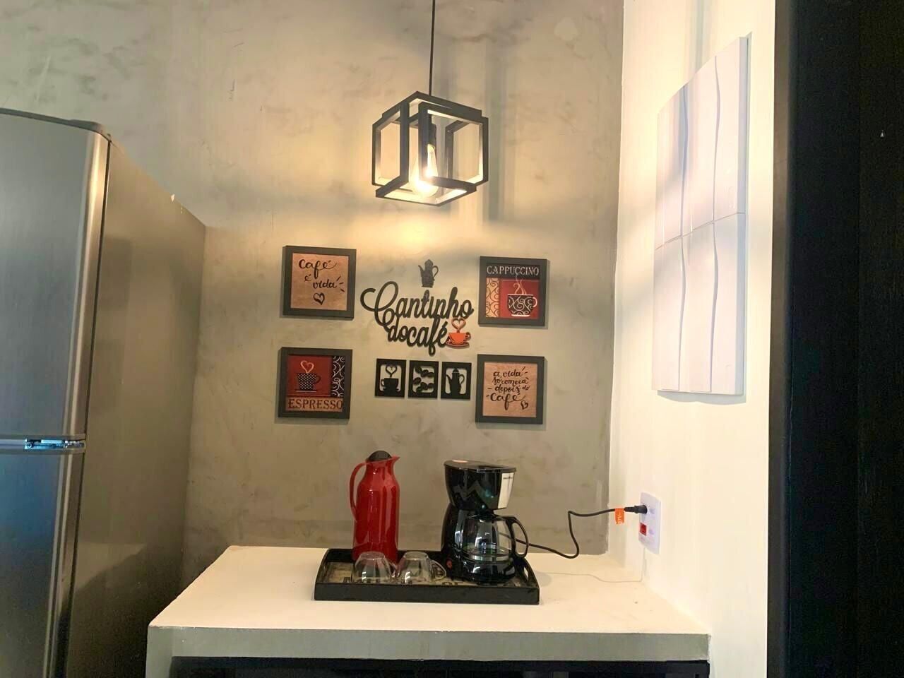 Cozinha com cafeteira, jarra vermelha e coador de café na bancada. Quadros emoldurados na parede e luminária pendente.