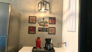 A kitchen with a coffee maker, a red pitcher, and a coffee pot on the counter. There are framed pictures on the wall and a hanging light fixture. - CASA PREMIUM - ARRAIAL DO CABO - RJ (Arraial do Cabo)