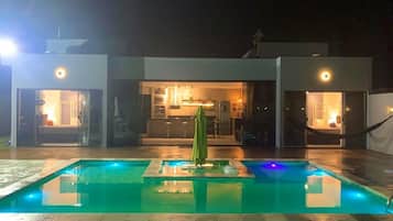 Uma piscina com um guarda-sol verde, um edifício moderno com portas de vidro e um interior bem iluminado.