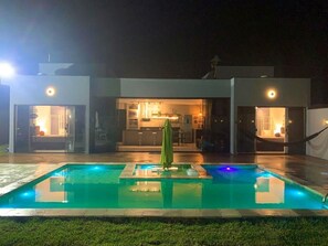 A swimming pool with a green umbrella, a modern building with glass doors, and a well-lit interior. - CASA PREMIUM - ARRAIAL DO CABO - RJ (Arraial do Cabo)