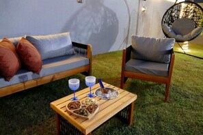 A cozy outdoor seating area with a sofa and a chair, a wooden table with snacks and drinks, and a decorative wall.