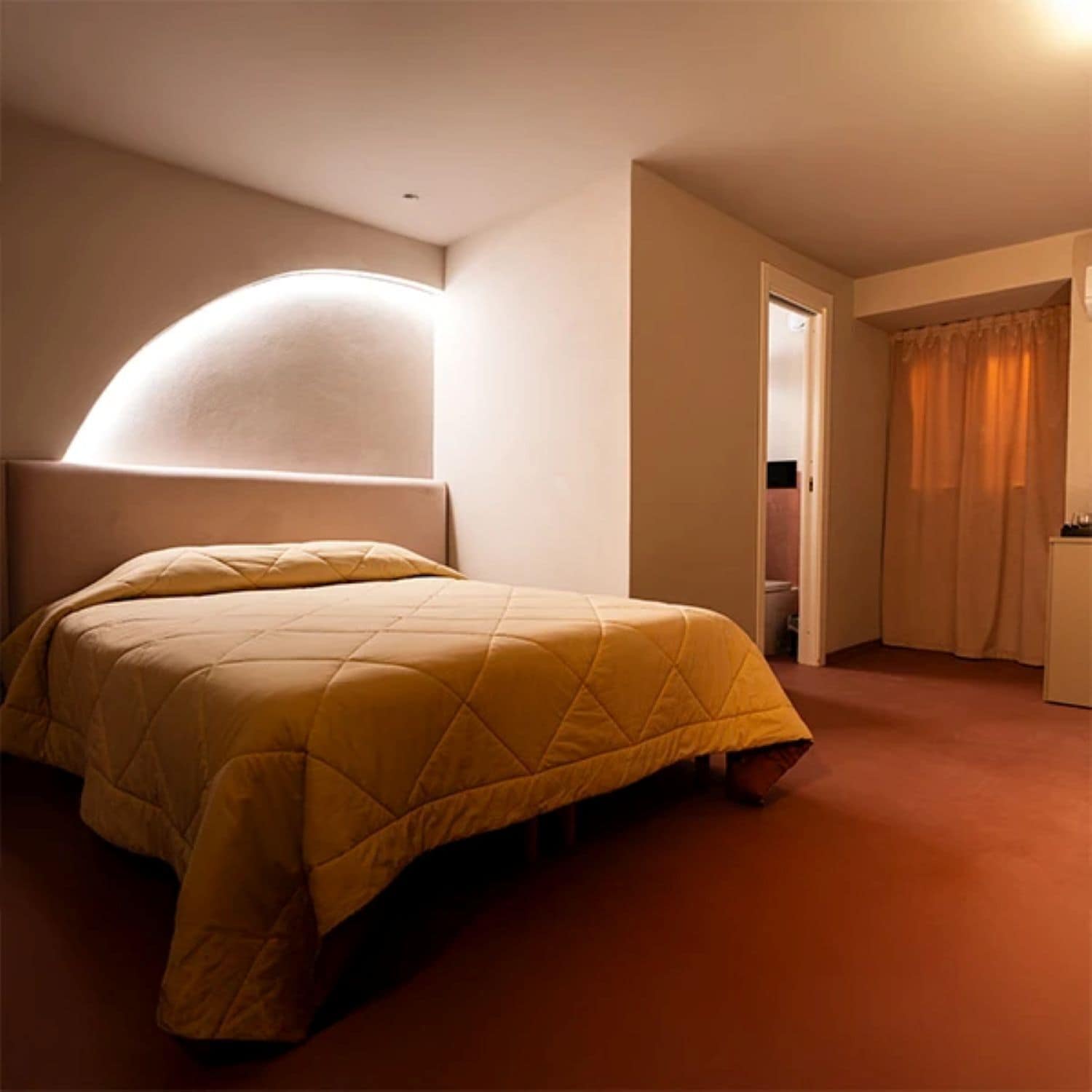 Un lit simple avec une tête de lit, un luminaire au plafond à la forme arrondie et une porte donnant sur une salle de bain.