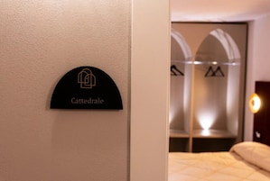 Ett hotellrum med en säng, en skylt som sitter på väggen och som visar namnet ”Cattedrale”, och en garderob med spegeldörrar.
