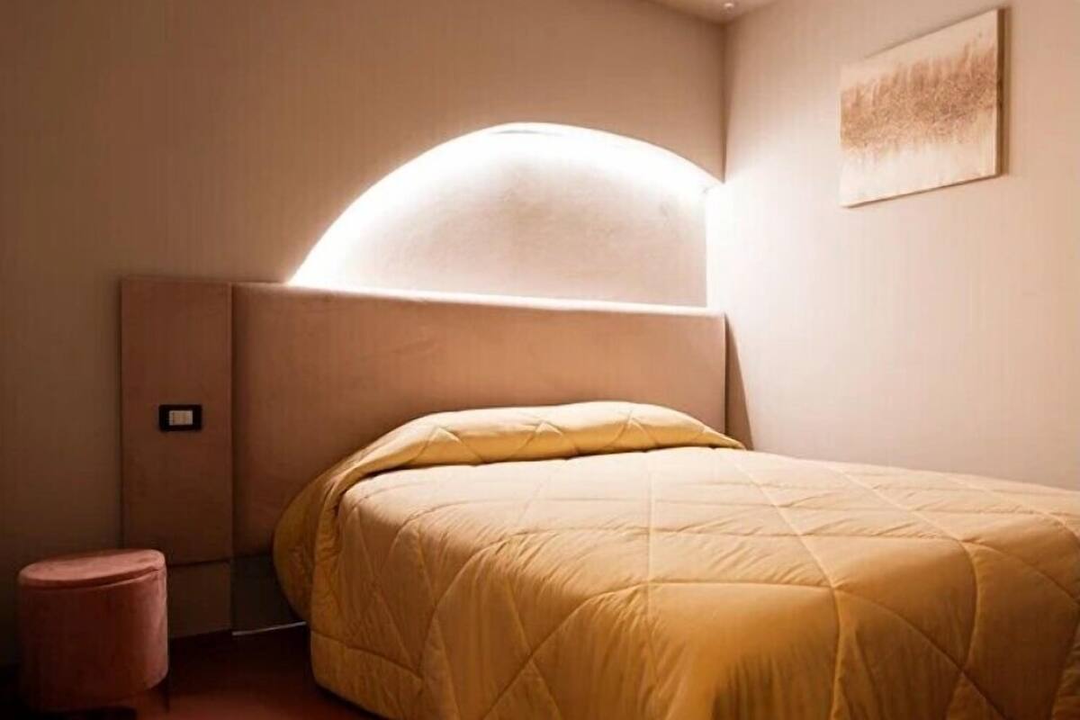 Ein Schlafzimmer mit einem Bett, einem Kopfteil, einem kleinen Hocker und einer Wandleuchte.