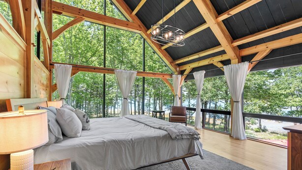 Une chambre spacieuse avec un grand lit, des poutres apparentes, un luminaire suspendu et une vue sur les arbres à travers de grandes fenêtres.