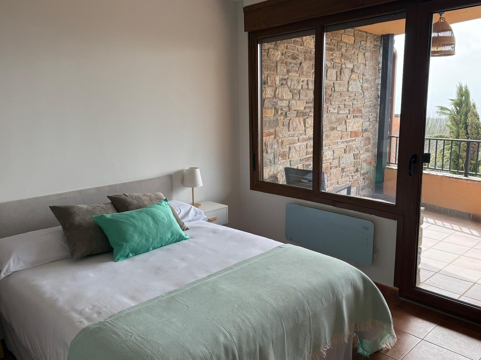 A bedroom with a bed, pillows, a bedside table with a lamp, and a view of a balcony with a stone wall and outdoor furniture.