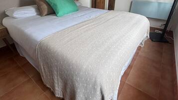 Een netjes opgemaakt bed met een bedsprei met patroon, een groen kussen en een wit kussen. Er is een houten deur en een aan de muur bevestigde televisie.