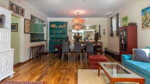 Uma sala de estar ampla com piso de madeira, uma mesa de centro de vidro, um sofá azul e uma área de jantar com um armário verde-água e um candelabro.