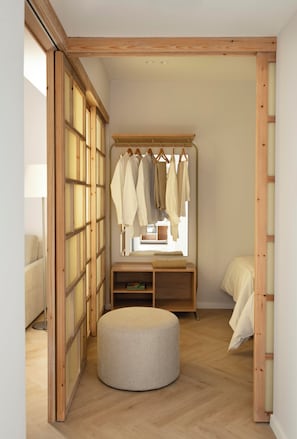 Een kamer met houten vloer, een rond poefje, een bed met witte lakens, een houten garderobe en een raam met natuurlijk licht.