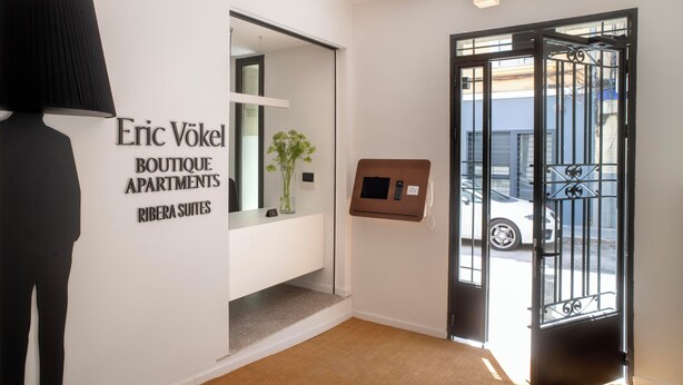Een modern interieur met een receptiebalie, een bord met Eric Vökel Boutique Apartments, een deur met een metalen hekwerk en een gang naar de ingang.