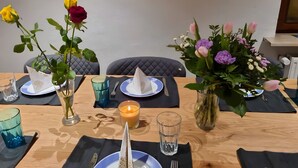 Mesa posta com pratos, talheres e uma vela, cercada por vasos com flores.