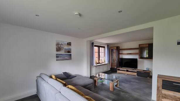 Uma sala de estar moderna com um sofá cinzento, uma mesa de centro de vidro e uma área de cozinha com armários de madeira e uma televisão.