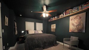 Een slaapkamer met een bed, een stoel, een plafondventilator en een boekenkast.
