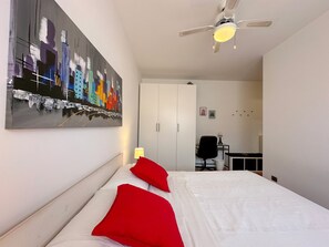 Une chambre à coucher avec un lit, des oreillers rouges, un ventilateur de plafond, un bureau avec une chaise et une armoire.