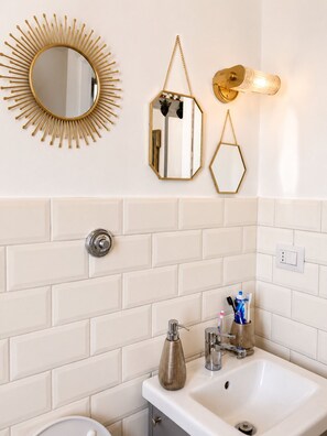 Un baño con pared de azulejos blancos, un espejo con diseño de sol dorado, un espejo hexagonal, una lámpara dorada, un lavamanos blanco y un porta cepillos de dientes.
