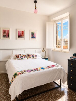 A bedroom with a bed, a dresser, a nightstand, a lamp, and a window with a view of buildings. - CASA FRIDA, cozy bright attic with terrace. (Roma)