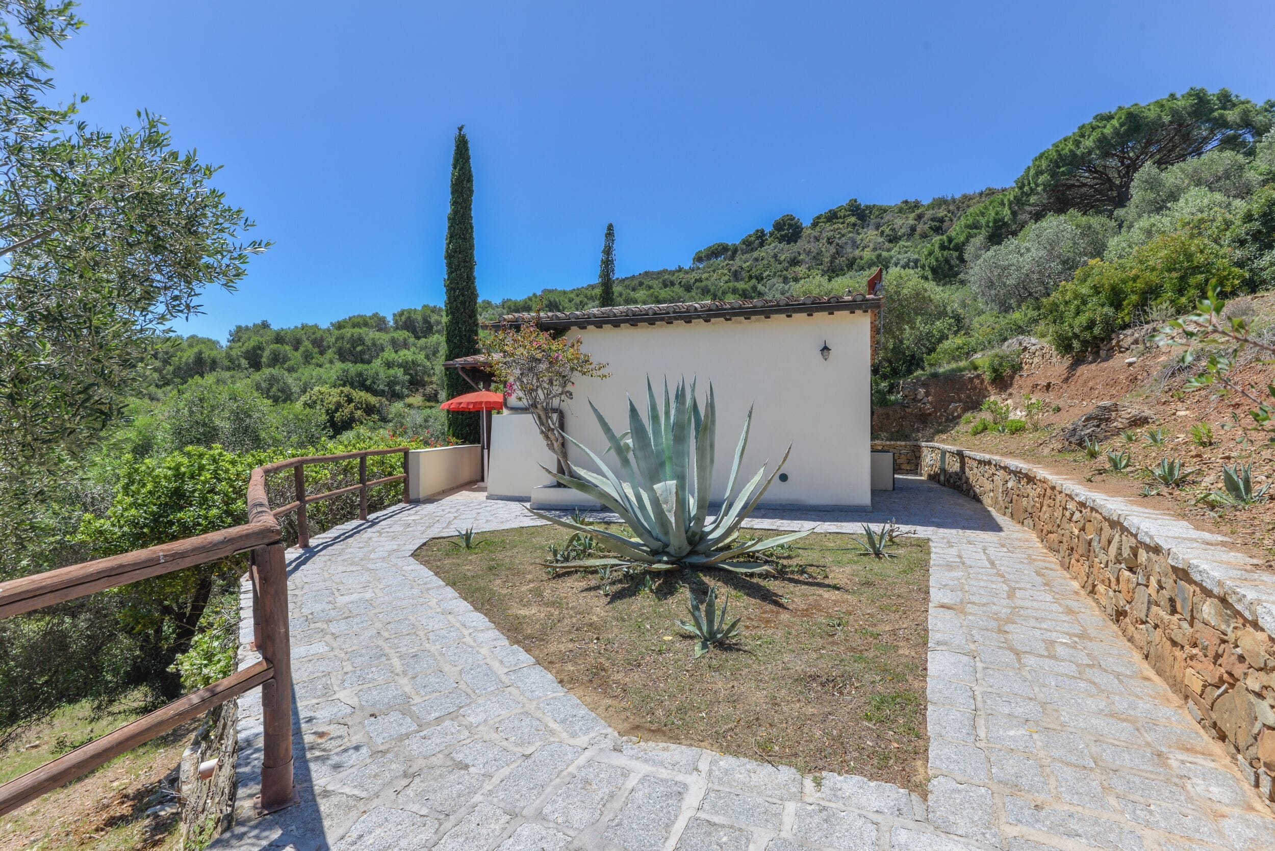 A stone pathway leading to a building with a small garden and a red umbrella.
