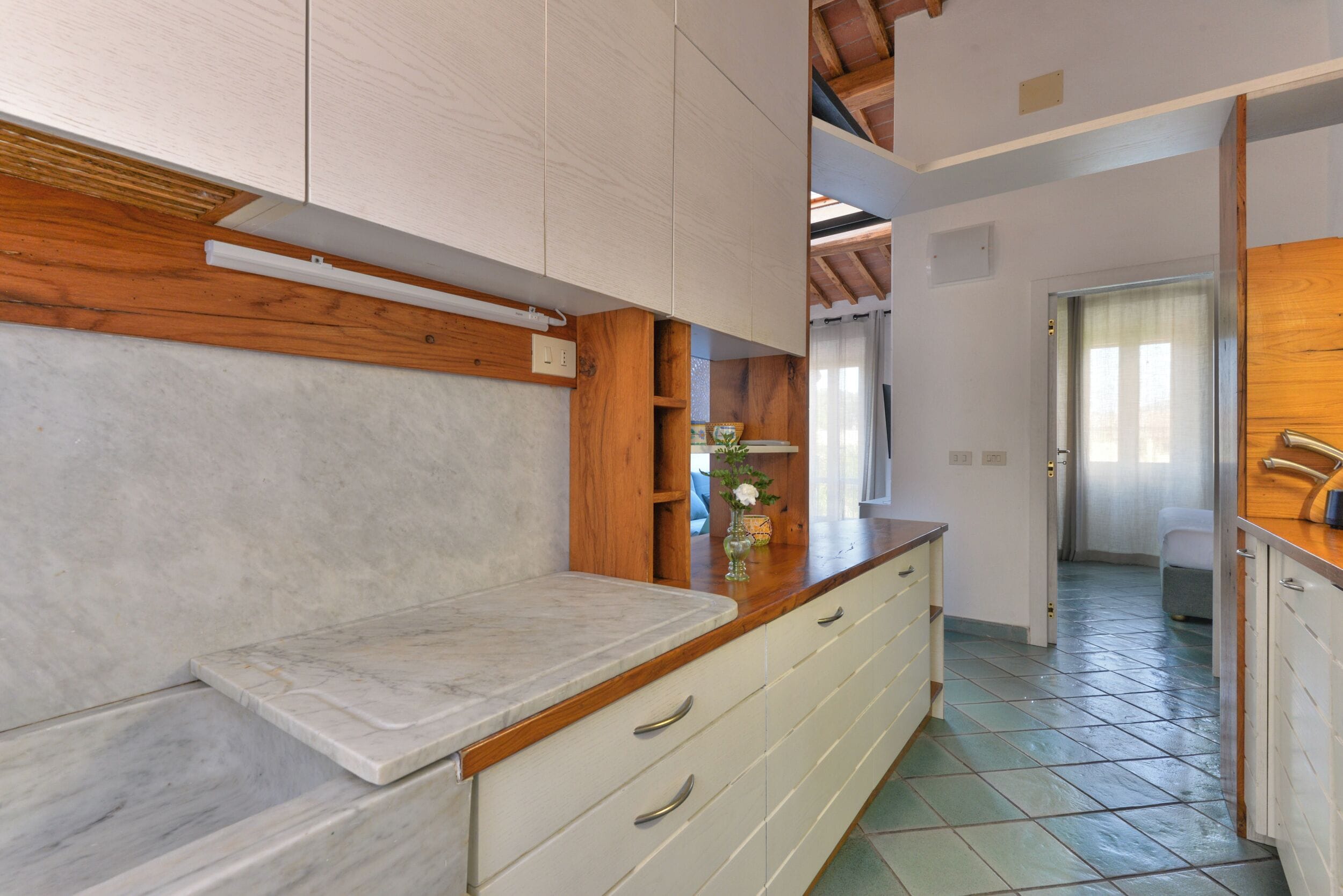A kitchen with white cabinets, a marble countertop, wooden beams, and a tiled floor.