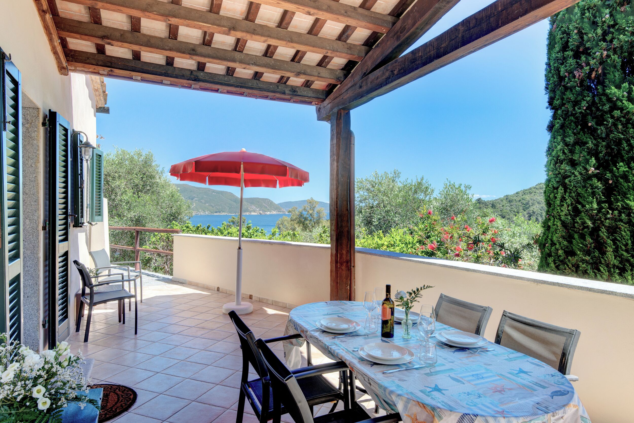 An outdoor dining area with a table set for two, a red umbrella, and a view of the sea and hills.