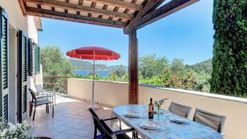 An outdoor dining area with a table set for two, a red umbrella, and a view of the sea and hills.