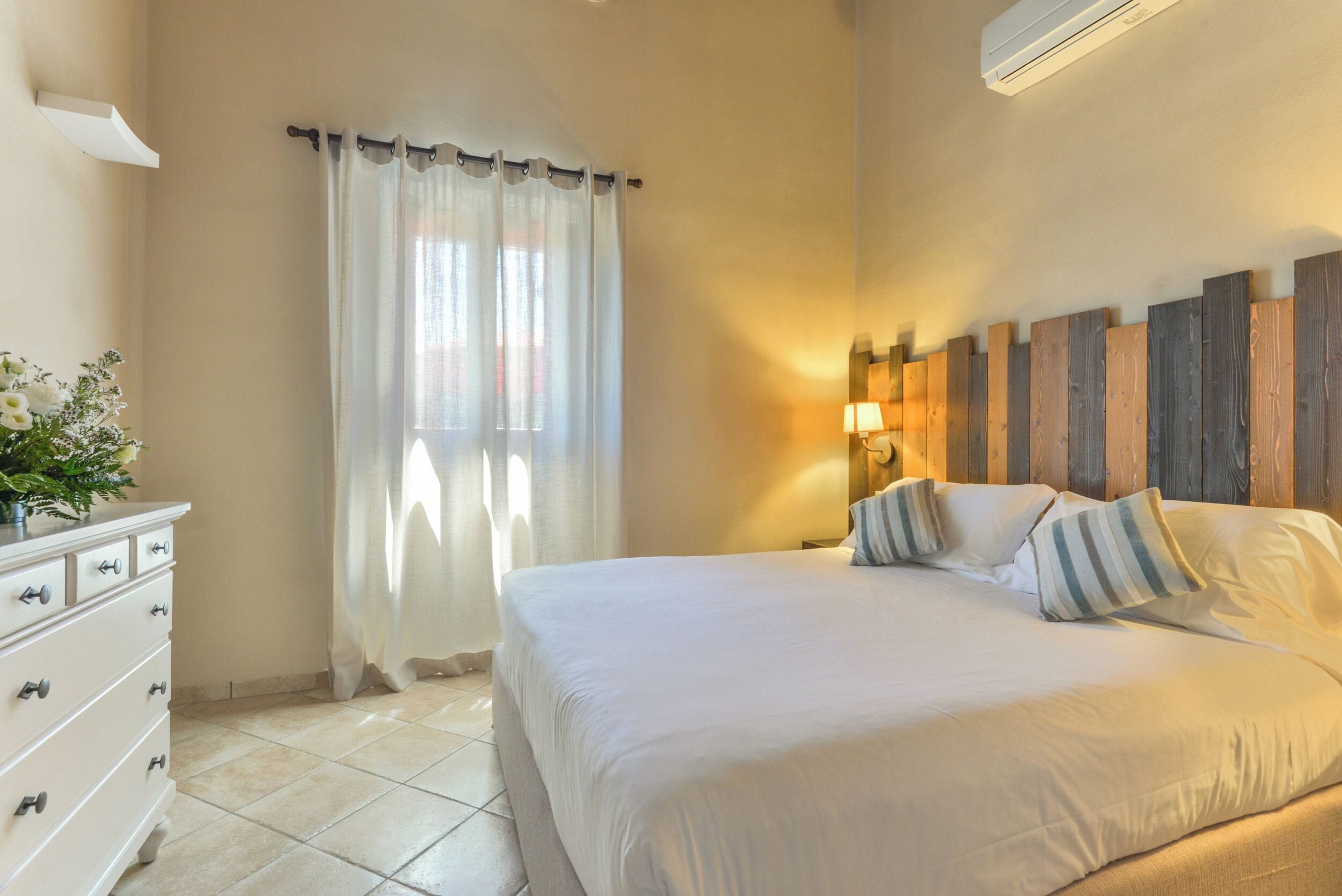 Une chambre à coucher comprenant un lit, une commode, une fenêtre avec des rideaux, une lampe fixée au mur et une tête de lit en bois.