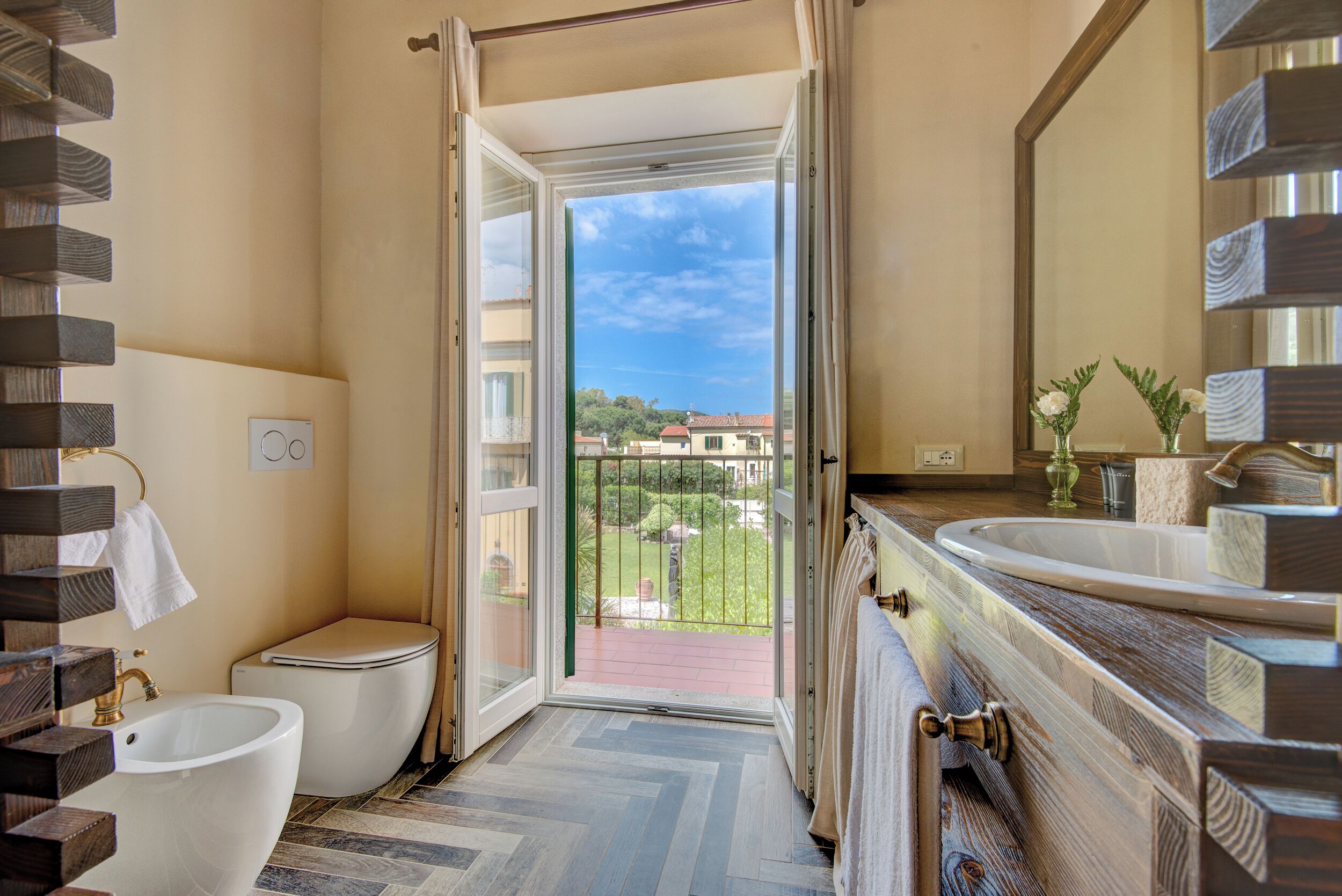 A bathroom with a toilet, sink, and open door leading to a balcony with a view of houses and greenery.