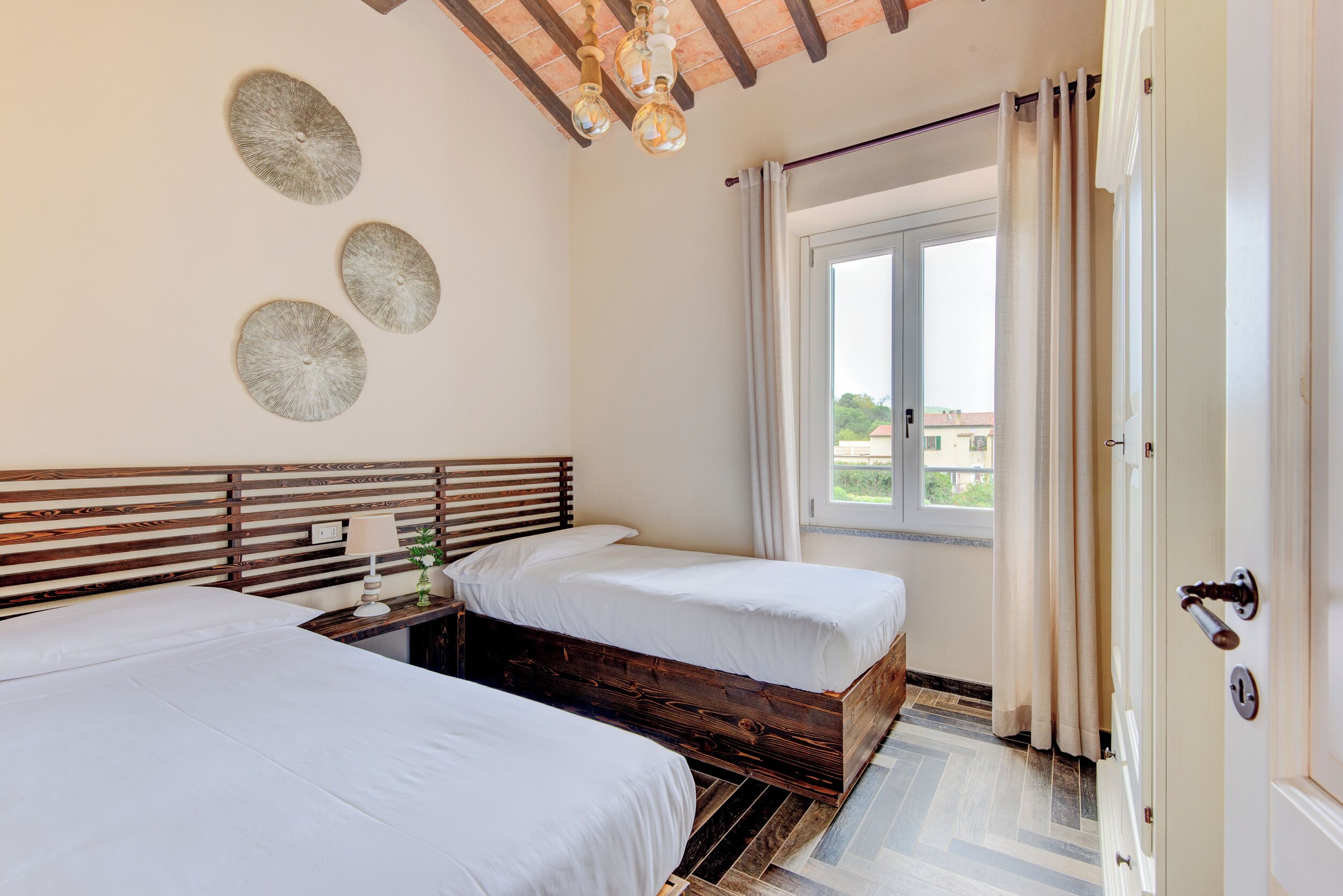 Un dormitorio con dos camas, cabeceros de madera, una mesita de noche con lámpara y una ventana con cortinas.