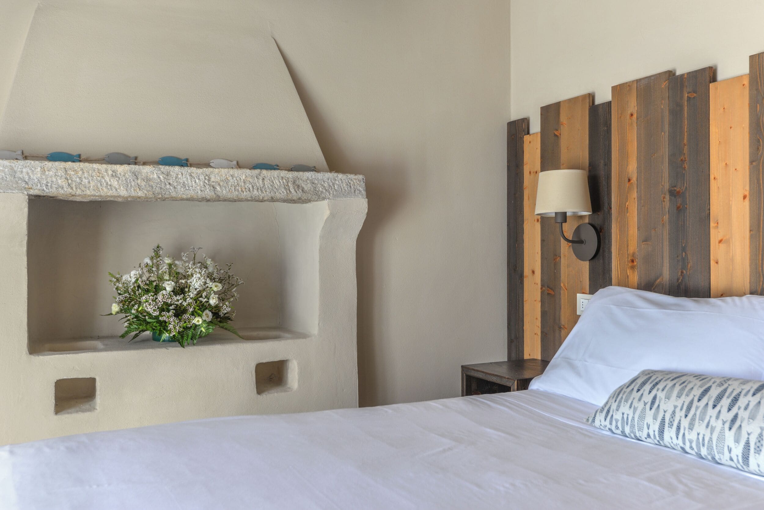 Ein Schlafzimmer mit einem Bett, einem Nachttisch, einer Wandlampe und einem Deko-Regal mit Blumen.