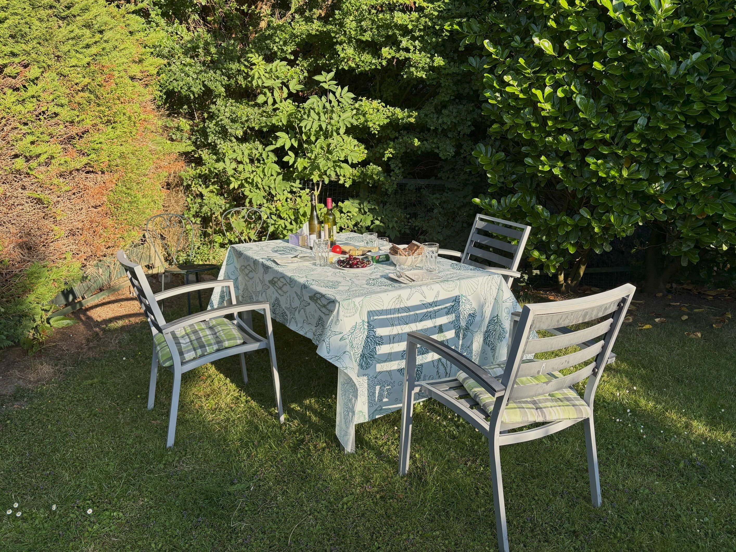 An outdoor dining table with a tablecloth, surrounded by chairs, set in a garden with greenery and trees.