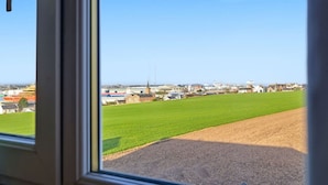 Blick durch ein Fenster auf eine ländliche Landschaft mit einem großen, grünen Feld, einer Kirche und mehreren Gebäuden in der Ferne.
