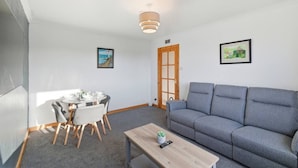 Ein kompakter Wohnraum mit einem Esstisch für vier Personen, einem grauen Sofa, einem Holztisch und einer Tür, die nach draußen führt.