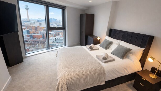 A modern bedroom with a large bed, a nightstand, a wardrobe, and a view of a cityscape through a large window. - Albert Dock Luxury Penthouse – Walk to Liverpool ONE (Liverpool)