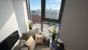 A modern living room with a large window, a grey sofa, a small side table, and a potted plant. - Albert Dock Luxury Penthouse – Walk to Liverpool ONE (Liverpool)
