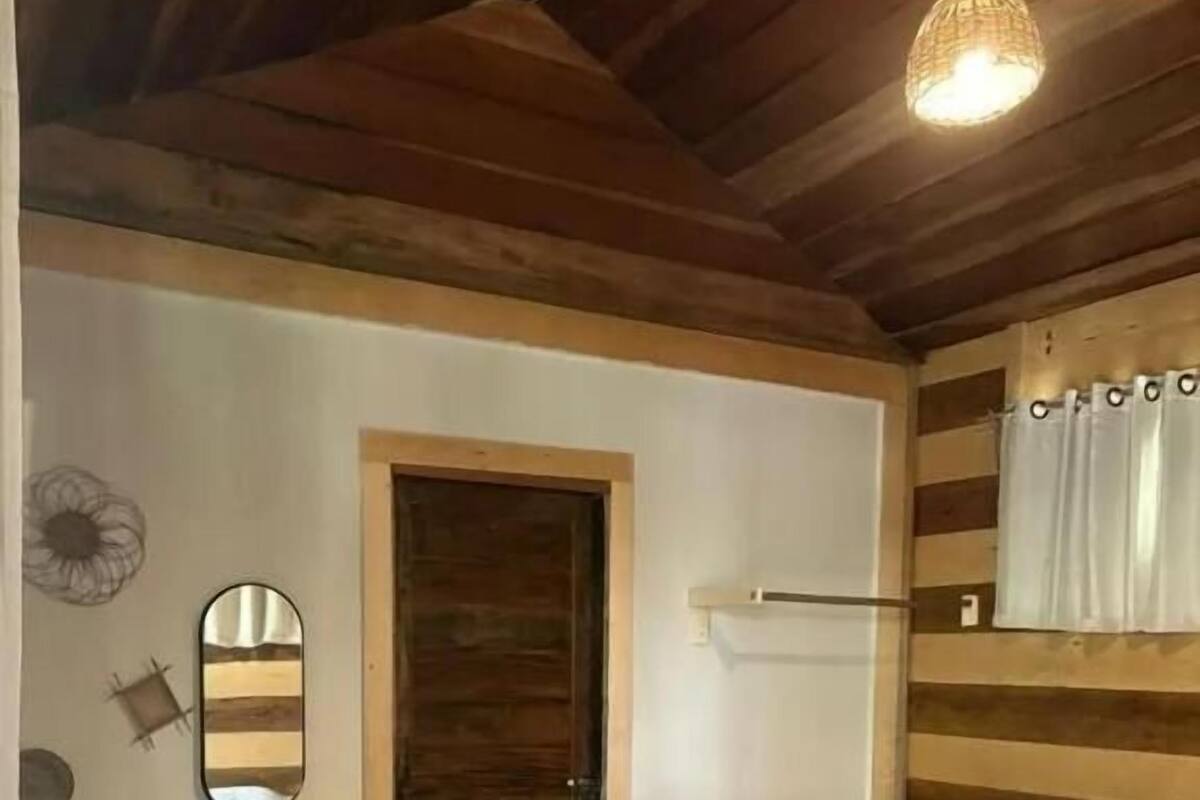 A room with wooden walls, a bed with a white bedspread, a small table, a mirror, and a hanging light fixture.