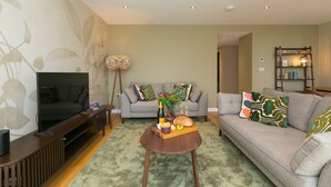 A living room with a TV, sofa, coffee table, and a bookshelf. - Karolva, St Ives - Ground Floor Apartment – On -Site Parking (St Ives)