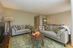A living room with two grey sofas, a wooden coffee table, a floor lamp, and a bookshelf. - Karolva-St Ives-Ground Floor Apartment-Parking (St Ives)
