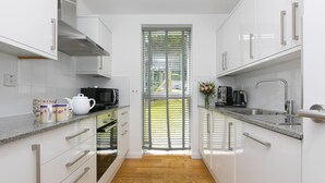 A modern kitchen with white cabinets, a granite countertop, a built-in oven, and a window with blinds. - Karolva, St Ives - Ground Floor Apartment – On -Site Parking (St Ives)