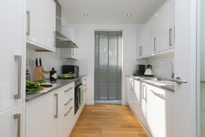 A narrow kitchen with white cabinets, a wooden floor, and a window with blinds. - Karolva-St Ives-Ground Floor Apartment-Parking (St Ives)