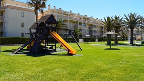 En lekplats med en rutschkana och en klätterställning, omgiven av en välskött gräsmatta och palmer, med en hotellbyggnad i bakgrunden.