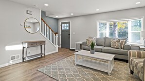 A modern living room with a staircase, a grey sofa, a coffee table, and a large window with blinds.